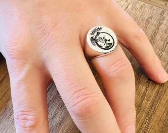 Può includere: Un anello a sigillo d'argento con un designo in bianco e nero di un uccello con una corona sulla testa.