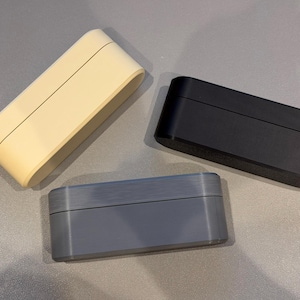 May include: Three rectangular boxes in cream, black, and gray. Each box has a rounded top and bottom, with a seam running along the center. The boxes are arranged on a light gray surface.