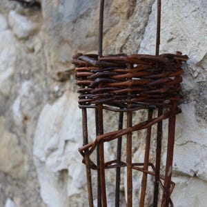 May include: A brown wicker basket hanging from a metal hook on a stone wall. The basket is made of woven twigs and has a cylindrical shape.
