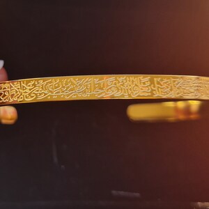 May include: A gold bangle bracelet featuring intricate Arabic calligraphy engraved on the surface.