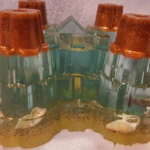May include: A clear resin castle with four towers, each topped with a copper-colored dome. The castle is filled with sand and seashells, creating a beach-themed design.