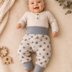 Könnte beinhalten: Ein Baby in einem cremefarbenen Langarmshirt mit Holzknöpfen und einer grauen Hose. Die Hose hat einen hellbeigen Hintergrund mit einem Muster aus blauen Elefanten. Das Baby liegt auf einer beigen Decke.