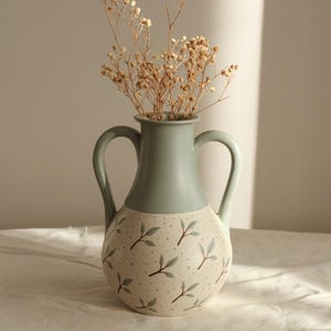 Könnte beinhalten: Eine Keramikvase mit zwei Henkeln, zweifarbig bemalt. Der obere Teil ist in einem gedämpften Grün gehalten, während der untere Teil cremefarben mit einem Blattmuster versehen ist. Trockenblumen sind in der Vase arrangiert.