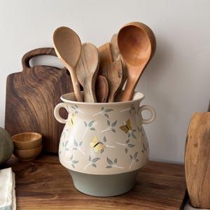 May include: A beige ceramic utensil holder with a green base, decorated with a leaf and butterfly pattern. It holds wooden spoons and ladles. A wooden chopping board and small wooden bowls are in the background.