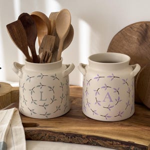 May include: Two cream-coloured ceramic utensil pots with handles, decorated with leaf wreaths and the letters "L" and "A". One contains wooden spoons. The pots are on a wooden board, suitable for a kitchen.