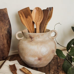 May include: A beige ceramic vase with two handles, filled with wooden kitchen utensils, including a spoon, spatula, and a fork. The vase is sitting on a wooden surface with a green plant in the background.