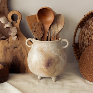 May include: A white ceramic pot with three legs and two handles, filled with wooden spoons and spatulas. The pot is sitting on a white linen tablecloth, next to a wooden chopping board and a wicker basket.