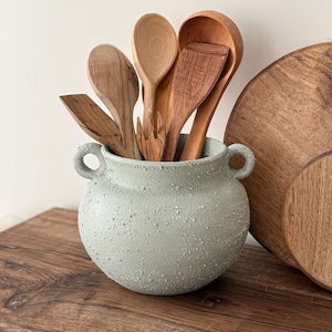 May include: A selection of wooden kitchen utensils, including spoons and spatulas, in a textured, pale green ceramic pot with handles. A wooden chopping board is visible, creating a rustic kitchen theme.