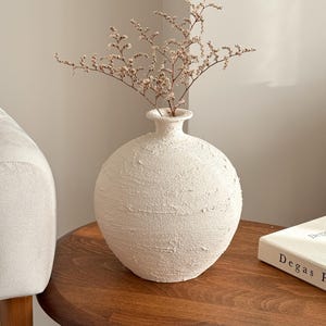 May include: A textured, cream-coloured ceramic vase with a rounded body and a slender neck, containing dried beige flowers. The vase is on a dark wooden side table next to a book titled "Degas Paintings".