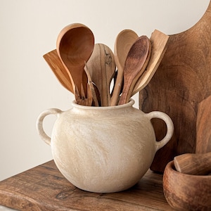 Könnte beinhalten: Eine Sammlung von Küchenutensilien aus Holz, darunter Löffel und Spatel, in einem cremefarbenen Keramiktopf mit Griffen. Ein Holzschneidebrett und ein Mörser mit Stößel sind ebenfalls zu sehen, was ein rustikales Küchenthema ergibt.
