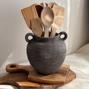 May include: A dark grey, textured ceramic jar with two side handles, filled with various light brown wooden kitchen utensils including spoons and spatulas, resting on two stacked wooden cutting boards with visible natural wood grain patterns.