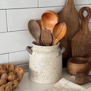 Op de afbeelding: Een gespikkelde keramische keukengerei houder met grijze handvatten bevat houten lepels en een pollepel. Houten snijplanken en een kleine houten kom staan op de achtergrond. Een mand met walnoten en een gestreepte doek zijn ook zichtbaar.