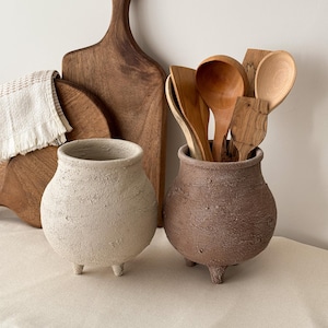 May include: Two small, textured ceramic pots with three legs each, one cream-colored and the other brown, sit on a neutral surface. Wooden spoons and cutting boards are in the background, suggesting a kitchen or dining theme.