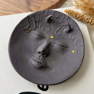 May include: Black ceramic incense holder shaped like a face with closed eyes and horns. The face has three yellow accents on the forehead and one on the cheek.