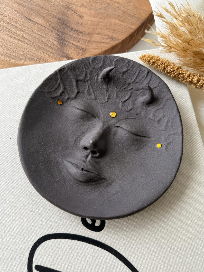 May include: Black ceramic incense holder shaped like a face with closed eyes and horns. The face has three yellow accents on the forehead and one on the cheek.