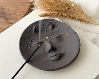 May include: A black ceramic incense holder shaped like a face with closed eyes and a small nose. The holder has gold accents around the eyes and on the forehead. A black incense stick is burning in the holder.