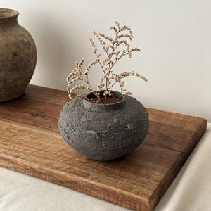 May include: A small, textured, dark gray ceramic vase with a round shape, holding dried beige flowers. The vase sits on a wooden tray, with another similar vase in the background. The overall aesthetic is rustic and minimalist.