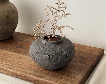 May include: A small, textured, dark gray ceramic vase with a round shape, holding dried beige flowers. The vase sits on a wooden tray, with another similar vase in the background. The overall aesthetic is rustic and minimalist.