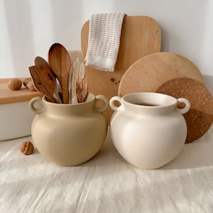 Op de afbeelding: Twee keramische vazen, één beige en één crème, met lusvormige handvatten. De beige vaas bevat houten keukengerei. Een houten snijplank, een kurken onderzetter en een patroonhanddoek zijn op de achtergrond.