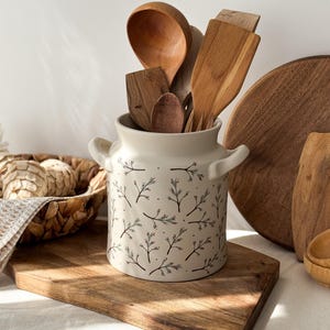 May include: A cream-coloured ceramic utensil holder with a floral pattern, filled with various wooden kitchen tools. The holder sits on a wooden chopping board, with a woven basket and additional wooden items in the background.