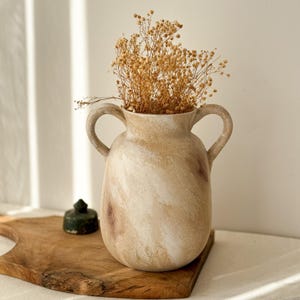 Könnte beinhalten: Eine beige Keramikvase mit zwei Henkeln, gefüllt mit getrockneten Blumen. Die Vase hat einen Marmoreffekt mit Braun- und Weißtönen. Ein kleiner grüner Gegenstand liegt auf einem Holzschneidebrett neben der Vase.
