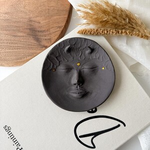 May include: A black ceramic dish with a face sculpted into the surface. The face has closed eyes, a nose, and a mouth. The dish has small gold dots around the face.