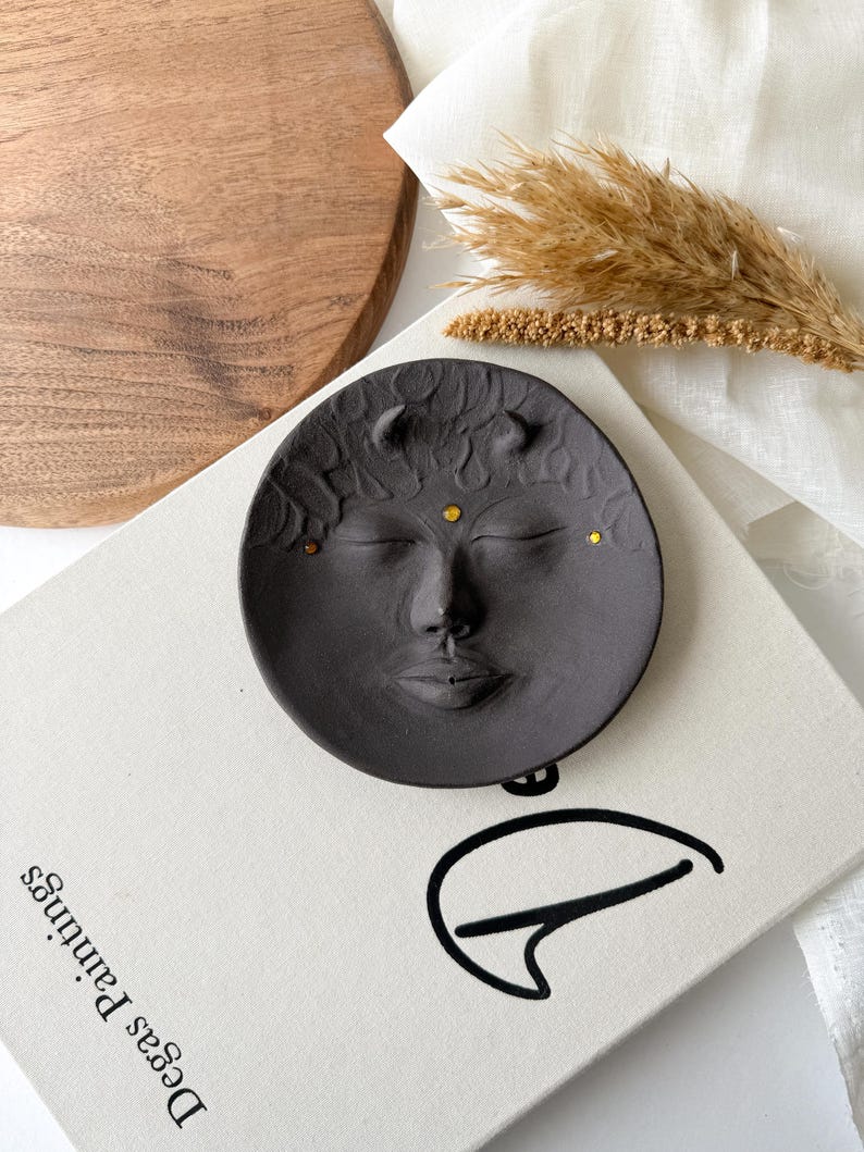 May include: A black ceramic dish with a face sculpted into the surface. The face has closed eyes, a nose, and a mouth. The dish has small gold dots around the face.