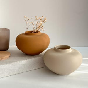 May include: Two ceramic vases, one in a light beige colour and one in a burnt orange colour. The orange vase is filled with dried flowers.