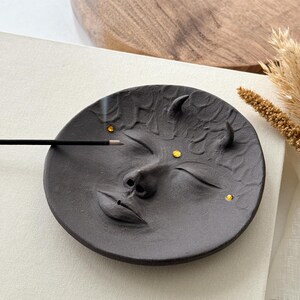 May include: Black ceramic incense holder shaped like a face with closed eyes and a small nose. The holder has three small gold accents on the face.