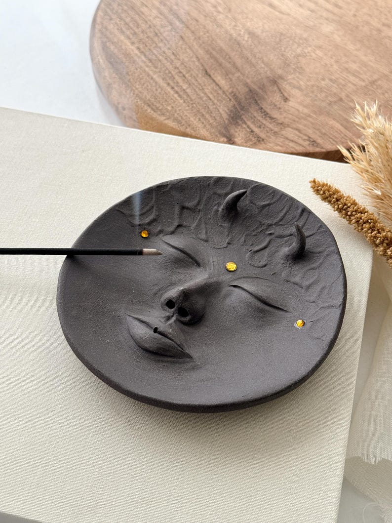 May include: Black ceramic incense holder shaped like a face with closed eyes and a small nose. The holder has three small gold accents on the face.