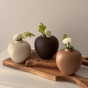 May include: Three small, spherical vases in cream, dark brown, and peach hues, each displaying a small arrangement of white flowers and green foliage. The vases are placed on a wooden serving board, offering a minimalist and natural aesthetic.