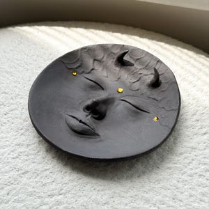 May include: A black ceramic dish with a face sculpted into the surface. The face has closed eyes, a nose, and a mouth. There are three small gold dots on the face, representing eyes and a third eye.