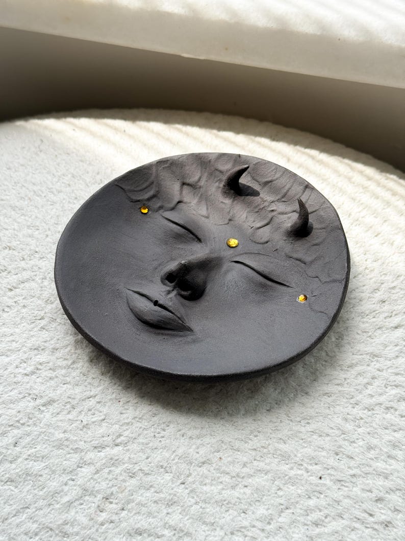 May include: A black ceramic dish with a face sculpted into the surface. The face has closed eyes, a nose, and a mouth. There are three small gold dots on the face, representing eyes and a third eye.