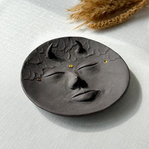 May include: A black ceramic dish with a face sculpted into the surface. The face has closed eyes, a nose, and a mouth. There are three small gold dots on the face.