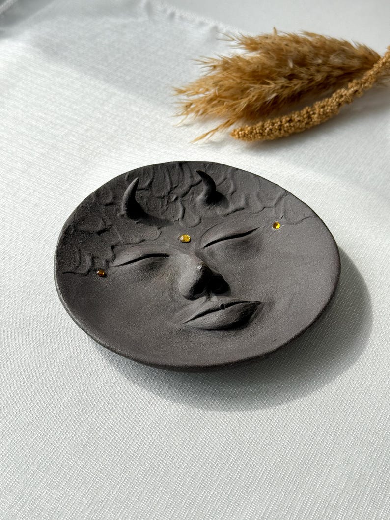 May include: A black ceramic dish with a face sculpted into the surface. The face has closed eyes, a nose, and a mouth. There are three small gold dots on the face.