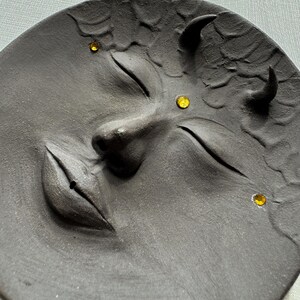 May include: A black ceramic plate with a face sculpted into it. The face has closed eyes, a slightly parted mouth, and two horns. There are three yellow rhinestones on the face.