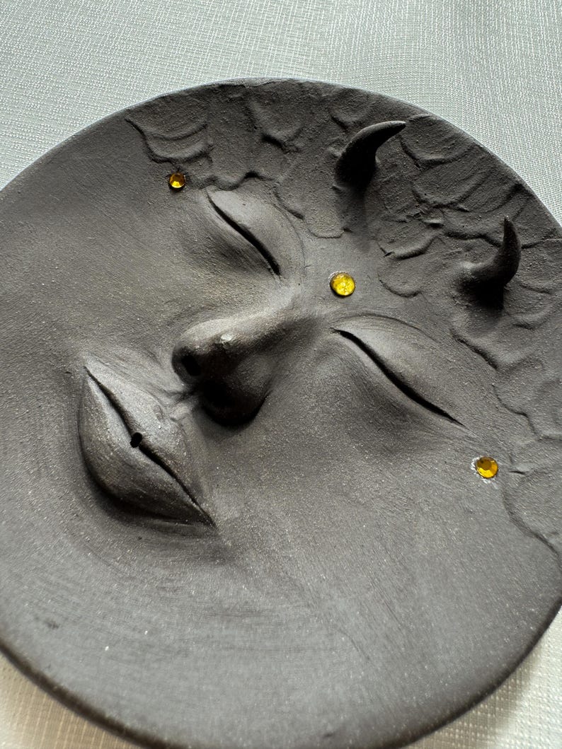 May include: A black ceramic plate with a face sculpted into it. The face has closed eyes, a slightly parted mouth, and two horns. There are three yellow rhinestones on the face.