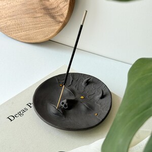 May include: Black ceramic incense holder shaped like a face with gold accents. The holder is on a book with the text "Degas P" visible. A black incense stick is in the holder.