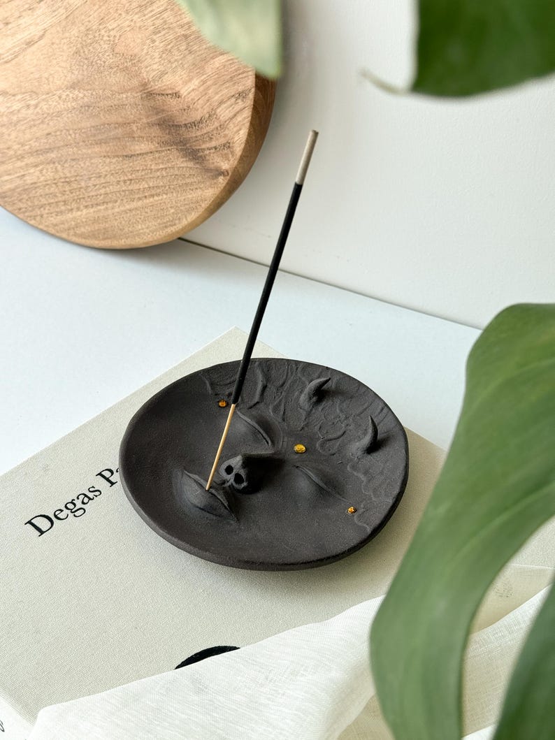 May include: Black ceramic incense holder shaped like a face with gold accents. The holder is on a book with the text "Degas P" visible. A black incense stick is in the holder.