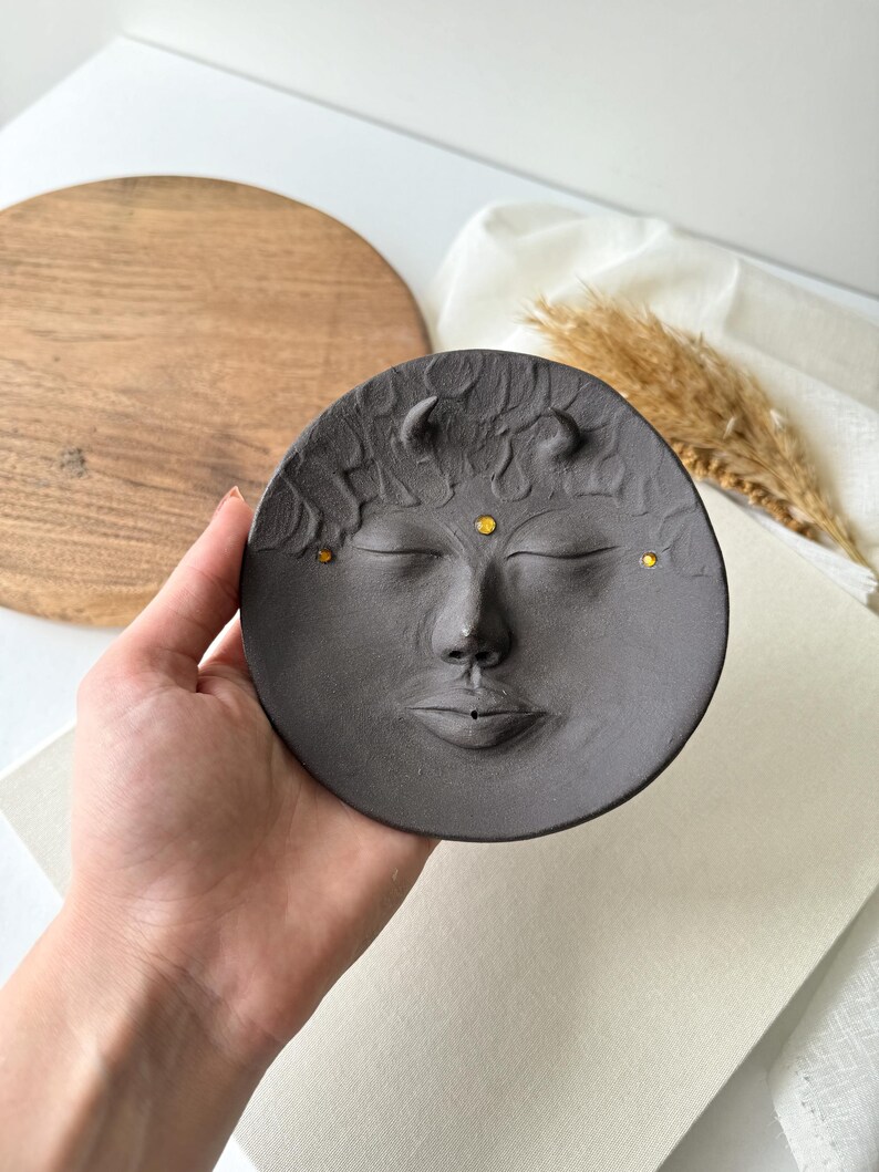 May include: A dark grey ceramic incense burner shaped like a face with closed eyes, a nose, and a mouth. The face has two small horns on the top and four gold dots on the forehead.