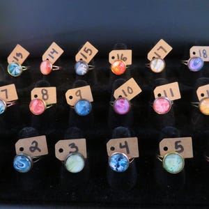May include: A display of rings with colorful, swirled designs. Each ring has a round, glass-like stone set in a copper-colored band. Small tags with numbers are attached to each ring, indicating different styles and sizes.