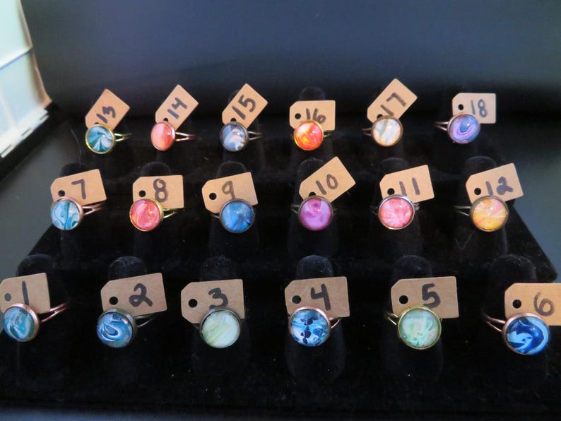 May include: A display of rings with colorful, swirled designs. Each ring has a round, glass-like stone set in a copper-colored band. Small tags with numbers are attached to each ring, indicating different styles and sizes.