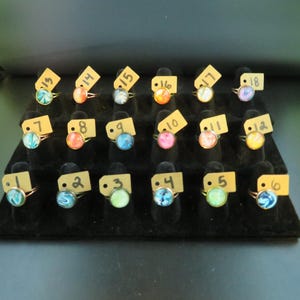 May include: A display of rings with colorful, swirled designs on a black velvet surface. Each ring has a gold-colored band and a round, glass-like stone. The rings are arranged in rows and labeled with small tags.