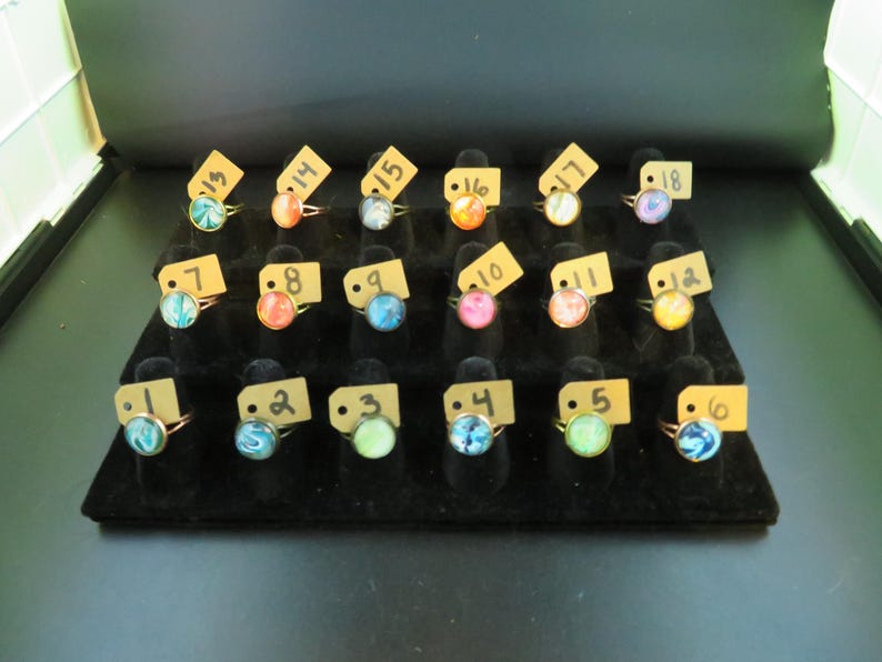 May include: A display of rings with colorful, swirled designs on a black velvet surface. Each ring has a gold-colored band and a round, glass-like stone. The rings are arranged in rows and labeled with small tags.
