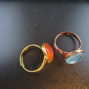 May include: Two adjustable rings with oval-shaped cabochon stones. One ring has a gold-tone band and an orange stone. The other ring has a copper-tone band and a blue stone. The rings are displayed on a dark surface.