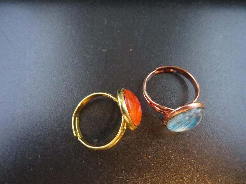 May include: Two adjustable rings with oval-shaped cabochon stones. One ring has a gold-tone band and an orange stone. The other ring has a copper-tone band and a blue stone. The rings are displayed on a dark surface.