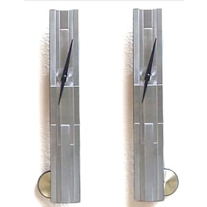 May include: Two vertical, silver-toned wall clocks with black hands. Each clock has a rectangular design with a pendulum at the bottom. The clocks are mounted on a light-colored wall.