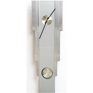 May include: A vertical, rectangular wall clock with a brushed silver finish. The clock features a circular, gold-toned clock face and a matching lower accent. Black hands indicate the time. The clock's design is modern and minimalist.