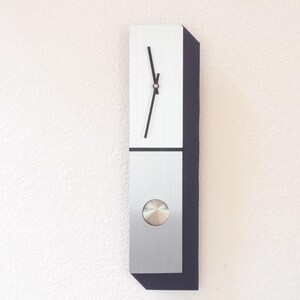 May include: A vertical rectangular wall clock with a brushed silver metal face and a dark side panel. The upper section features two thin black hands, while the lower section contains a circular silver pendulum disc centered on the metal surface.