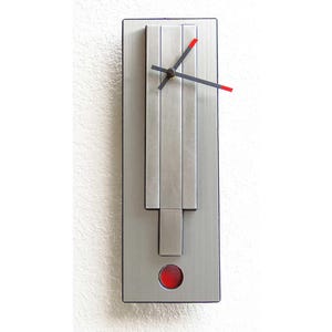 May include: A rectangular, silver-toned wall clock with a modern design. The clock features black hands with red tips, a red circular accent at the bottom, and a brushed metal finish. The clock's dimensions are approximately 18 inches in height and 6 inches in width.